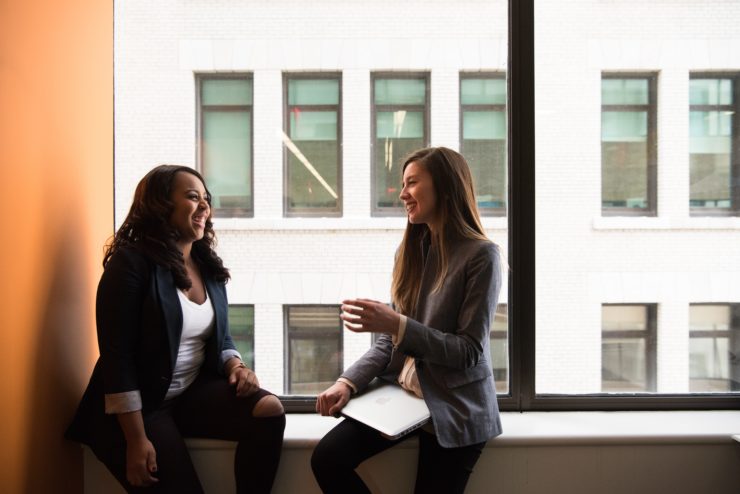 Photo of two women at work, happily chatting while sitting against the office windows.