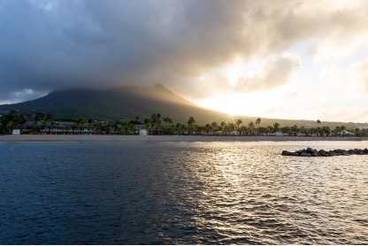 An image of the ocean in the foreground with palm trees and a mountain in the background. The sun is setting behind the right side of the mountain. 