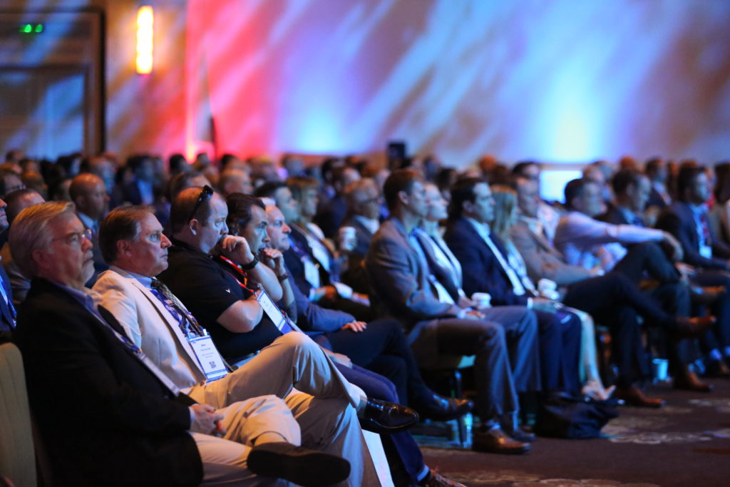 Large audience looks towards stage with colorful lights shining on the ballroom wall