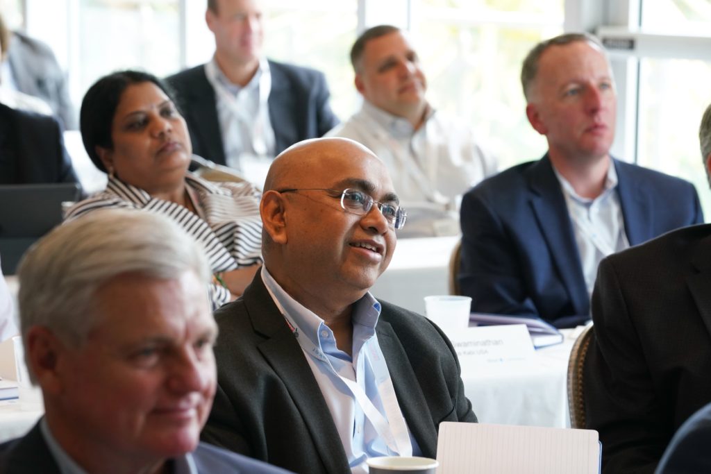 Attendees at classroom tables in a well lit room face the stage