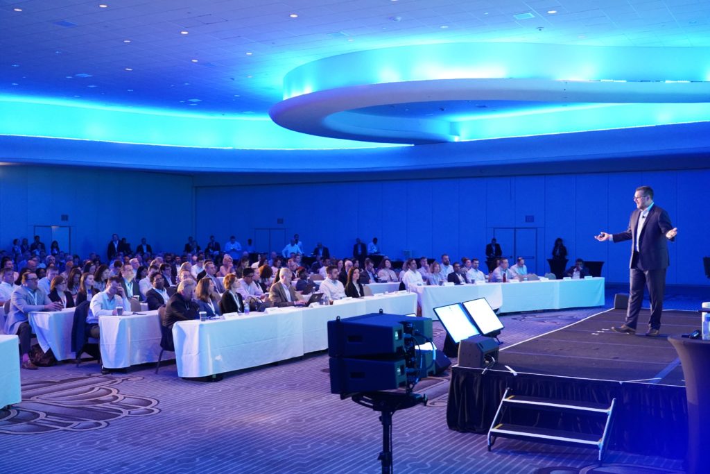 Speaker addresses room of attendees at classroom tables in room with blue lighting