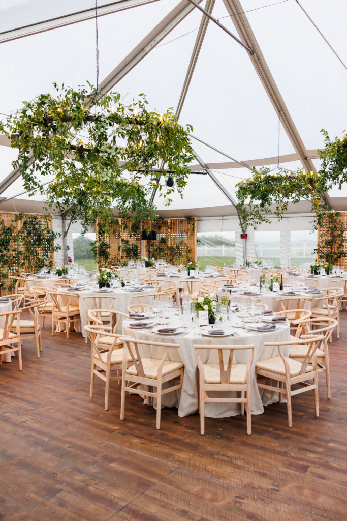 Evening event set up in a tent featuring neutral table décor and chandeliers dripping with greenery