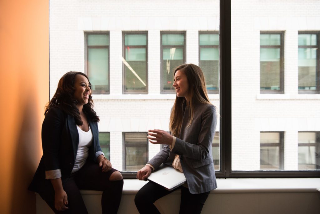 Photo of two women at work, happily chatting while sitting against the office windows. 